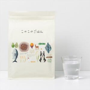 Cream-colored bag of dog food with colorful animal illustrations and Japanese text, placed beside a glass of water.